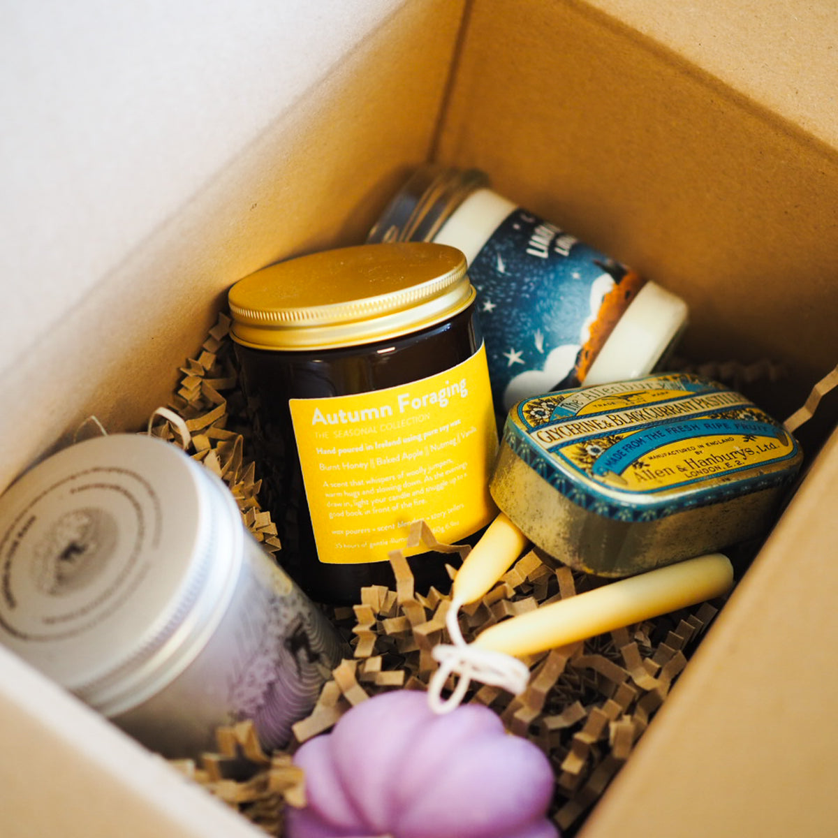 A curated gift box containing a dark glass candle jar labeled 'Autumn Forening' with a gold lid, a blue and gold tin candle, a purple flower-shaped candle, and other wrapped candles nestled in brown crinkle paper inside a cardboard box.