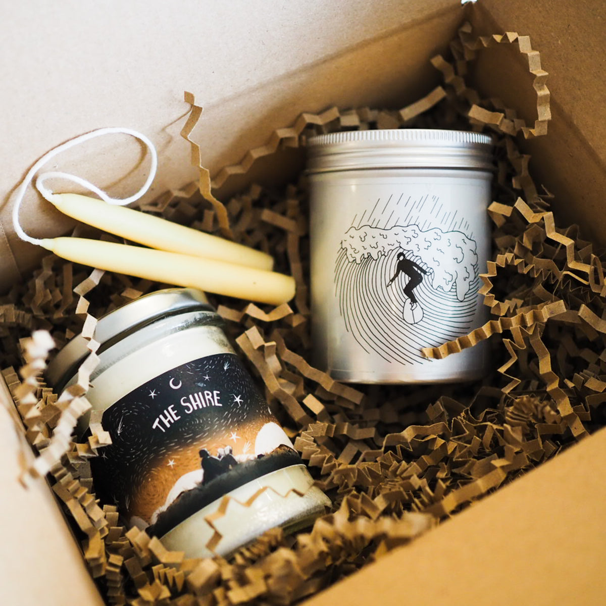 A gift box containing a silver tin candle with a wave surfer illustration, a jar candle labeled 'THE SHIRE' with a sunset graphic, and two yellow beeswax taper candles nestled in brown crinkle paper.