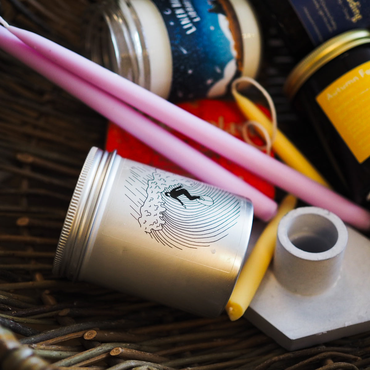 A close-up of a silver metal tin with a black surfboard graphic, lying on a wicker tray with pink and yellow candles, a glass jar, and a small white cylindrical object.